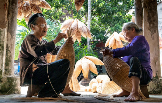 Deux artisans traditionnels (femmes) tressant des nasses de pêche en bambou (vannerie artisanale) dans un atelier extérieur, montrant un savoir-faire fait main et ancestral.